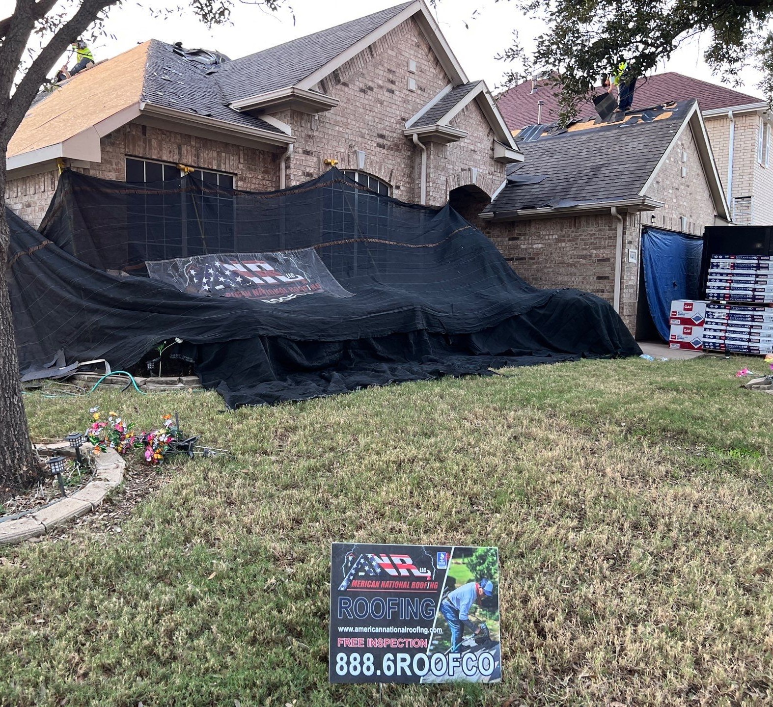 Roof replacement in progress in Keller 76244 with flower beds and windows covered for protection and an American National Roofing yard sign